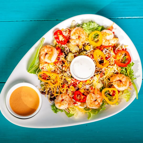 Shrimp salad with bell peppers, cheese, and two dressings on a white plate over a blue table.